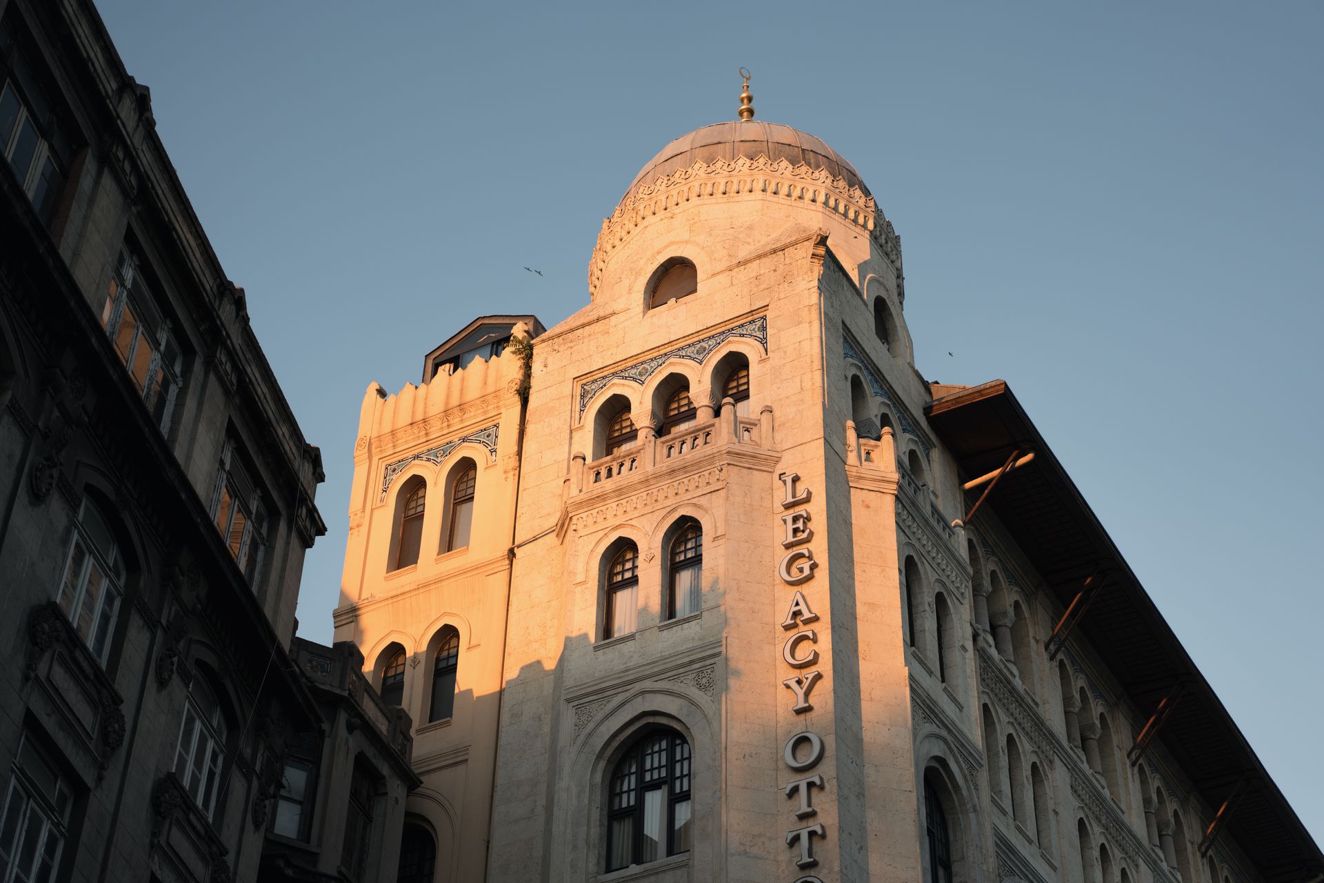 Majestic neoclassical facade and domed corner turrets of the historic Legacy Ottoman Hotel building bathed in golden afternoon sunlight.