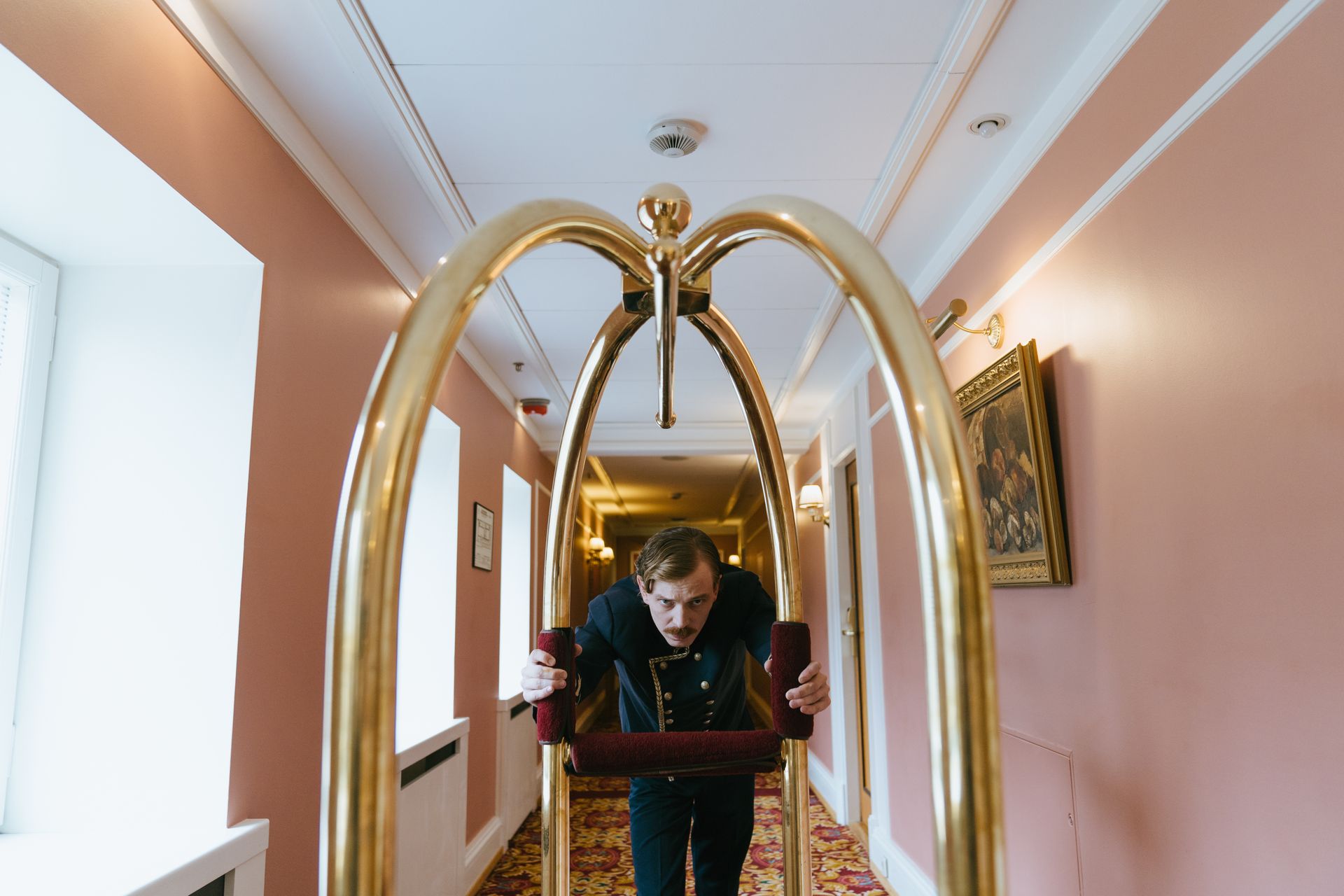 Uniformed hotel bellhop pushing a polished brass luggage cart down an elegant carpeted luxury hotel corridor.