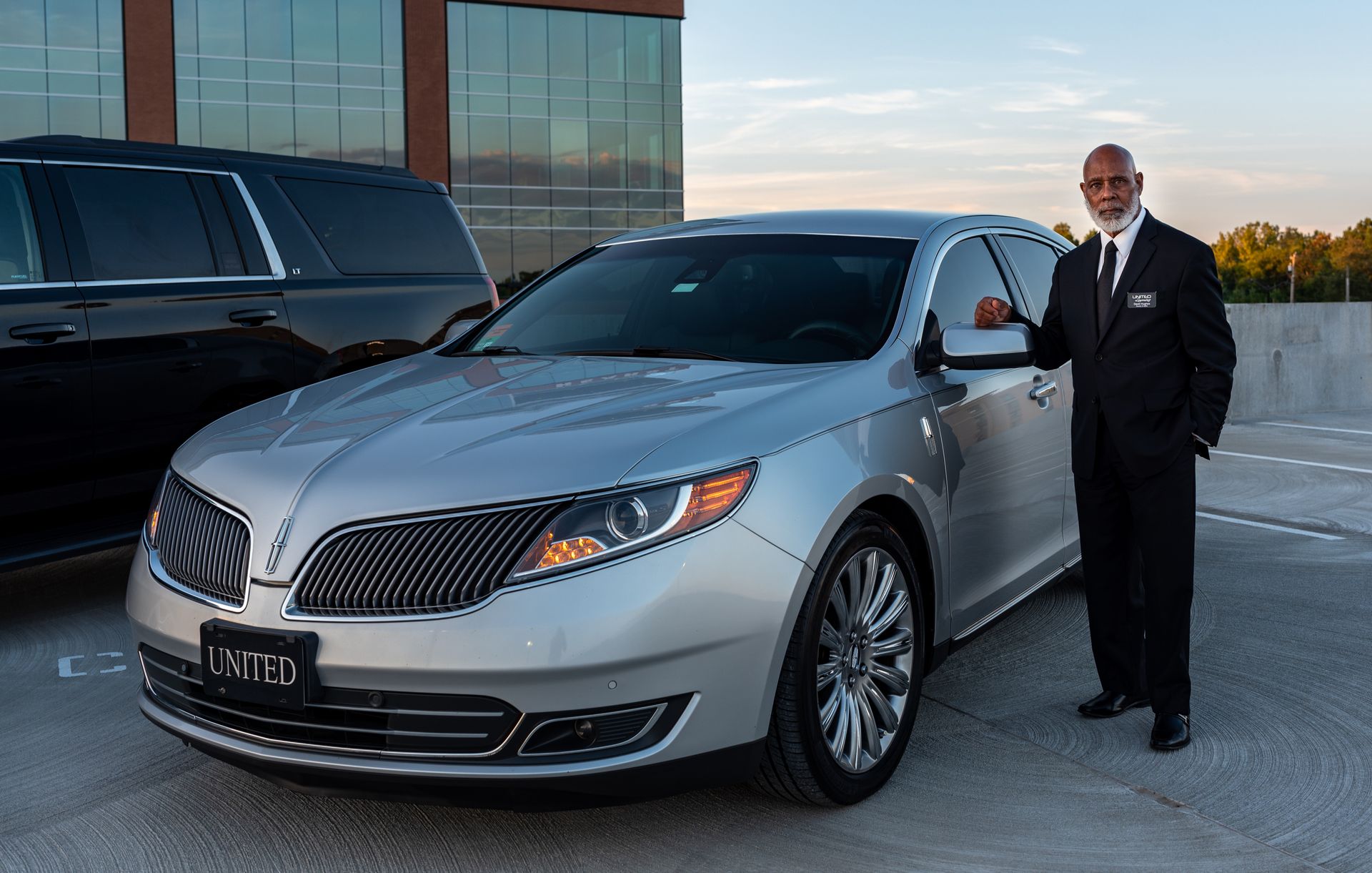 Professional chauffeur in a formal black suit standing attentively beside a premium executive transfer vehicle.