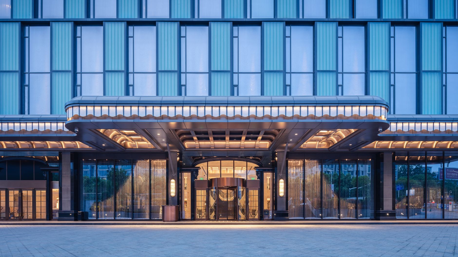 Modern urban corporate hotel entrance featuring a polished glass canopy and valet driveway.
